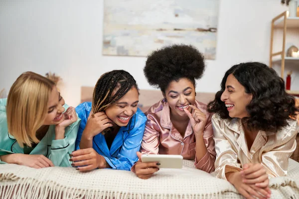Сміються і багатоетнічні подружки в барвистій піжамі розважаються, використовуючи смартфон разом, лежачи на ліжку під час вечірки піжами вдома, прив'язуючи час у зручному сні — стокове фото