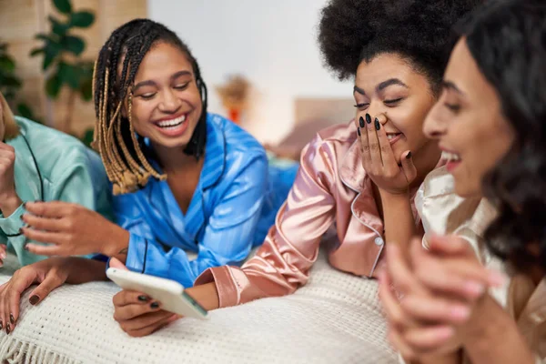 Fröhliche afrikanisch-amerikanische Frau, die Mund bedeckt und Smartphone benutzt, während sie mit multiethnischen Freundinnen im farbenfrohen Pyjama auf dem heimischen Bett Spaß hat, die Zeit in bequemer Nachtwäsche bindet — Stockfoto