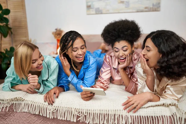 Позитивные многоэтнические женщины в красочной пижаме веселятся и используют смартфон и расслабляются на кровати во время пижамной вечеринки дома, время сближения в удобной спальной одежде — стоковое фото