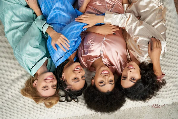 Draufsicht auf fröhliche multiethnische Freundinnen im bunten Pyjama, die sich umarmen und in die Kamera schauen, während sie zu Hause im Schlafanzug auf dem Bett liegen, Bindungszeit, Schlummerparty — Stockfoto