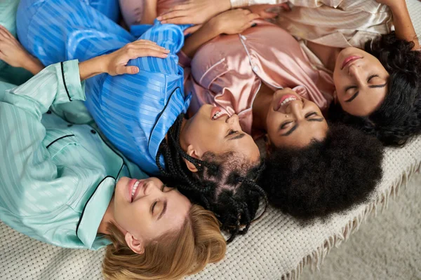 Top view of multiethnic girlfriends in colorful pajama laughing and hugging while lying on bed during pajama party in bedroom at home, bonding time in comfortable sleepwear, slumber party — Stock Photo