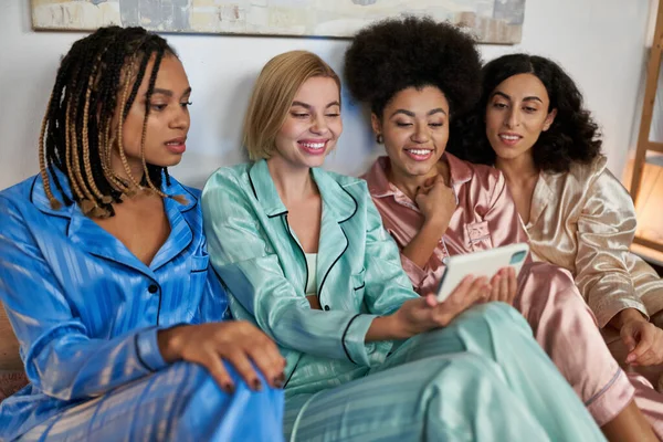 Mujer rubia sonriente usando y sosteniendo teléfono inteligente cerca de novias multiétnicas en pijama colorido durante la fiesta de pijama en casa, tiempo de unión en ropa de dormir cómoda, fiesta de pijama - foto de stock