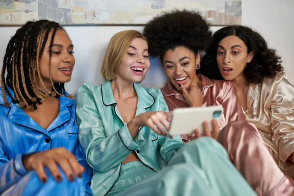 Excited blonde woman using blurred smartphone and sitting near cheerful multiethnic girlfriends in colorful pajama during girls night at home, bonding time in comfortable sleepwear, slumber party — Stock Photo