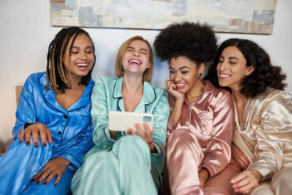Mujer rubia joven y riendo sosteniendo teléfono inteligente borroso cerca de novia multiétnica en pijama colorido durante la noche de las niñas mientras está sentado en la cama en casa, tiempo de unión en ropa de dormir cómoda - foto de stock