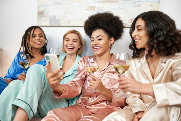 Alegre mujer afroamericana sosteniendo teléfono inteligente borroso cerca de novias multiétnicas en pijamas de colores sosteniendo copas de vino durante la noche de las niñas en casa, tiempo de vinculación, fiesta de pijamas - foto de stock