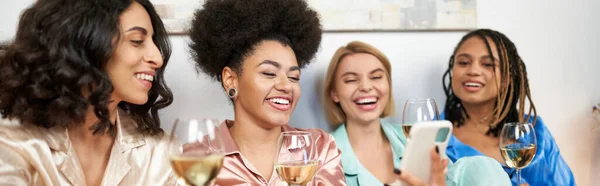 Sonriente mujer afroamericana usando un teléfono inteligente borroso cerca de novias multiétnicas en pijama colorido con gafas de esposa durante la noche de las niñas en casa, tiempo de vinculación en ropa de dormir cómoda, pancarta - foto de stock