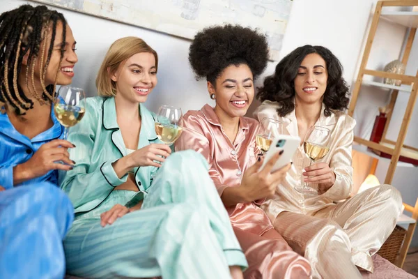 Novias positivas y multiétnicas en pijama colorido usando teléfono inteligente y sosteniendo vasos de vino mientras se sienta en la cama durante la noche de las niñas en casa, ropa de dormir cómoda, fiesta de pijamas - foto de stock