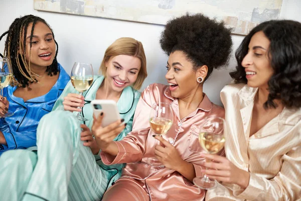 Smiling african american woman showing blurred smartphone at multiethnic girlfriends in colorful pajama during girls night with glasses of wine at home, bonding time in comfortable sleepwear — Stock Photo