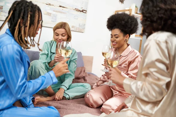 Sonriente mujer afroamericana sosteniendo un vaso de vino mientras habla con novias multiétnicas con copas de vino durante la noche de las niñas en el dormitorio en casa, tiempo de vinculación en ropa de dormir cómoda - foto de stock