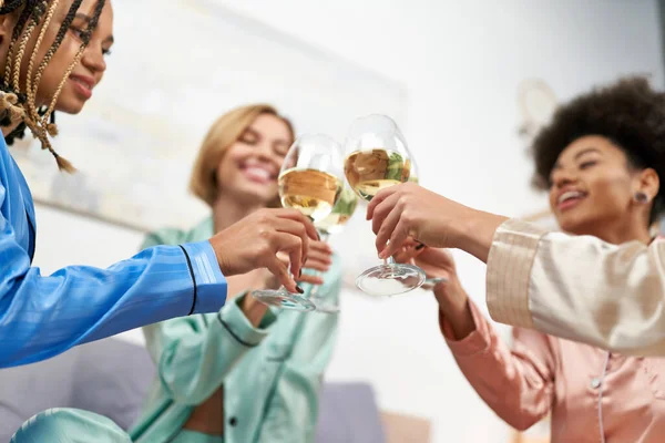 Low angle view of blurred and cheerful multiethnic girlfriends in colorful pajama toasting with wine during girls night at home, bonding time in comfortable sleepwear, slumber party — Stock Photo