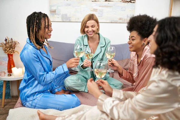 Copines positives et multiethniques en pyjama coloré tenant des verres de vin tout en parlant pendant la nuit des filles sur le lit à la maison, coller le temps dans des vêtements de nuit confortables, soirée pyjama — Photo de stock