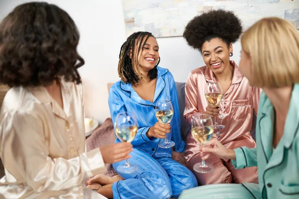 Positive and african american women in colorful pajama holding glasses of wine and talking to blurred girlfriends while sitting on bed during girls night at home, bonding time in sleepwear — Stock Photo