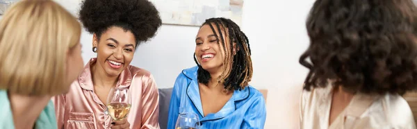 Улыбающиеся африканские американские женщины в красочной пижаме, держащие бокалы вина и разговаривающие с размытыми девушками во время девичника дома, время сближения в удобной сонной одежде, баннер — стоковое фото