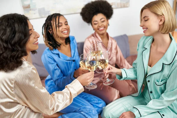 Mujeres multiétnicas sonrientes en coloridas tostadas de pijama con copas de vino con novias afroamericanas borrosas mientras están sentadas en la cama durante la fiesta de pijama, tiempo de unión en ropa de dormir cómoda - foto de stock