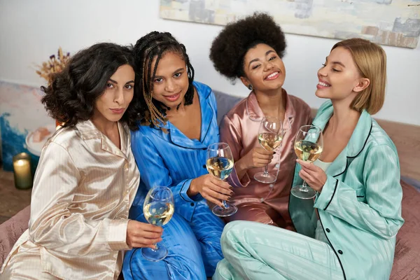 Портрет радостных и многонациональных подружек в красочной пижаме, держащих бокалы вина и смотрящих в камеру, сидящих на кровати во время пижамной вечеринки дома, связывающих время в удобной сонной одежде — стоковое фото