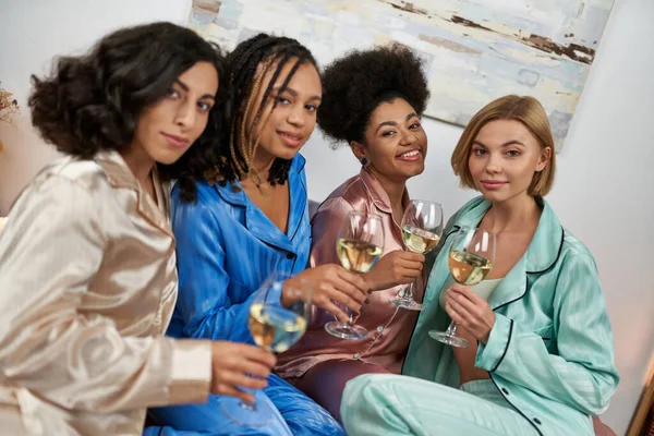 Sonrientes novias multiétnicas en pijamas coloridos sosteniendo copas de vino y mirando a la cámara durante la fiesta de chicas en casa, fiesta de pijamas, tiempo de unión en ropa de dormir cómoda - foto de stock