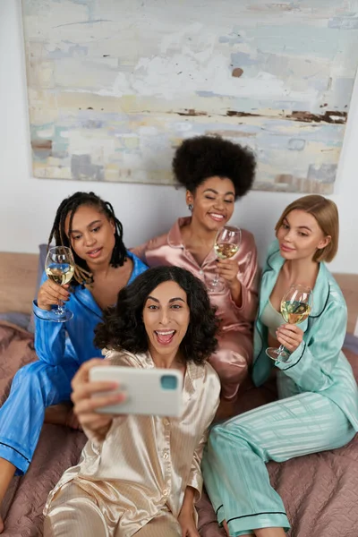 Високий кут зору веселих багатоетнічних подруг в барвистих піжамах тримає вино і приймає селфі на смартфон, сидячи на ліжку вдома, прив'язуючи час, партія слимаків — стокове фото