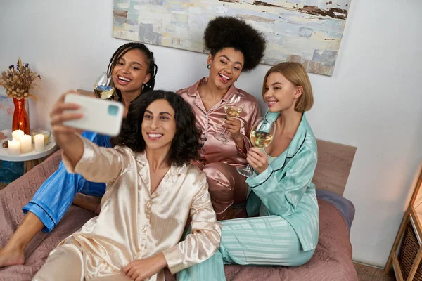 High angle view of multiracial woman in pajama taking selfie with cheerful multiethnic girlfriends with wine and sitting on bed at home, bonding time in comfortable sleepwear, slumber party — Stock Photo