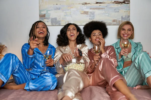 Веселі багатоетнічні подружки в барвистій піжамі тримають келихи вина і попкорну під час перегляду телевізора на ліжку під час дівчаток вночі, поєднуючи час у зручному сні — стокове фото