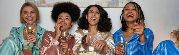 Lächelnde multikulturelle Freundinnen im bunten Pyjama mit Wein und Popcorn vor dem Fernseher während der Nacht der Mädchen zu Hause, Schlummerparty, Bindungszeit in bequemer Nachtwäsche, Banner — Stockfoto