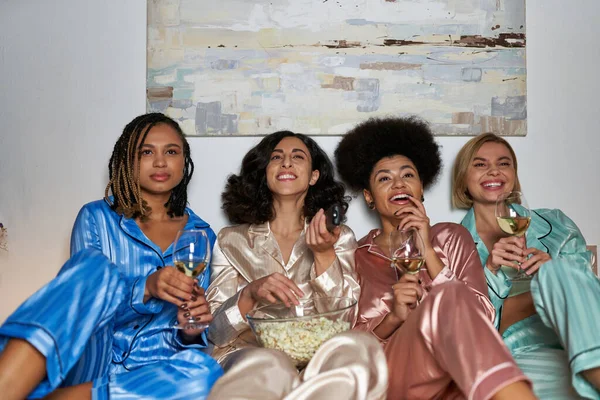 Улыбающиеся многорасовые женщины в пижаме, смотрящие телевизор и держащие попкорн и бокалы вина, сидящие на кровати во время девичника дома, проводящие время в удобной сонной одежде, пижамная вечеринка — стоковое фото