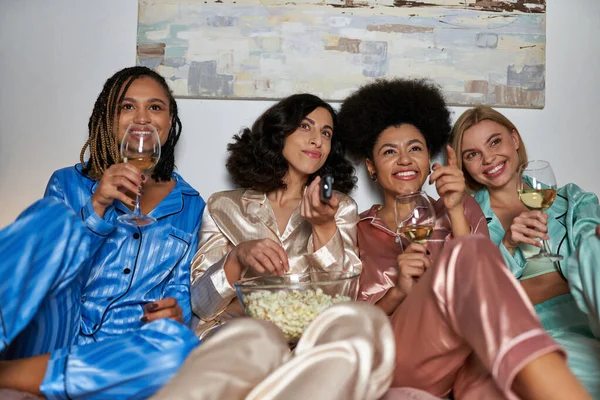 Alegres mujeres multiculturales en pijama sosteniendo copas de vino y palomitas de maíz mientras ven películas juntas en la cama durante la noche de las niñas en casa, tiempo de unión en ropa de dormir cómoda - foto de stock