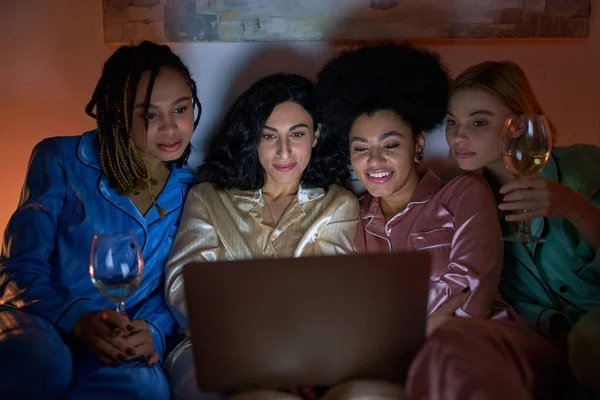 Lächelnde multiethnische Freundinnen im farbenfrohen Pyjama mit Weingläsern und verschwommenem Laptop während der Nacht der Mädchen zu Hause, Bindungszeit in bequemer Nachtwäsche, Schlummerparty — Stockfoto