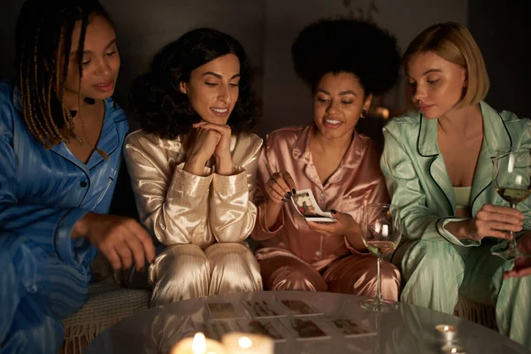 Lächelnde Afroamerikanerin mit Tarot-Karten neben multiethnischen Freundinnen mit Wein und Kerzen bei Pyjama-Party zu Hause, Bindungszeit in bequemer Nachtwäsche, Wahrsagerei — Stockfoto