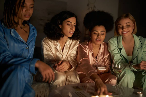 Lächelnde multiethnische Freundinnen im farbenfrohen Pyjama beim Betrachten von Tarotkarten auf dem Tisch neben einem Glas Wein und Kerzen in der Nacht zu Hause, Bindungszeit in bequemer Nachtwäsche, Schlummerparty — Stockfoto