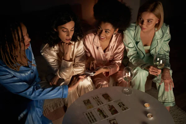 Vue aérienne de la femme afro-américaine en pyjama tenant des cartes de tarot près de copines multiethniques avec du vin près des bougies sur la table pendant la nuit des filles à la maison, le temps de liaison dans des vêtements de nuit confortables — Photo de stock