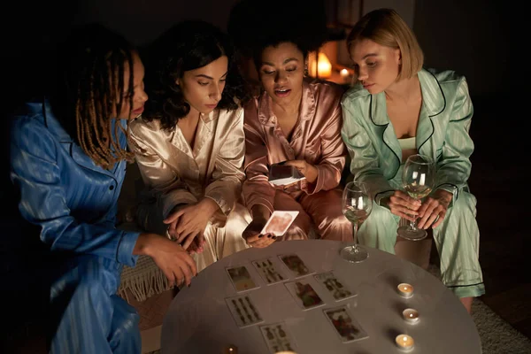 Гадание, африканская американка в пижаме держит карты Таро и принимает к многонациональным девушкам возле бокалов вина и свечей во время девочек ночь на дому, время связи — стоковое фото