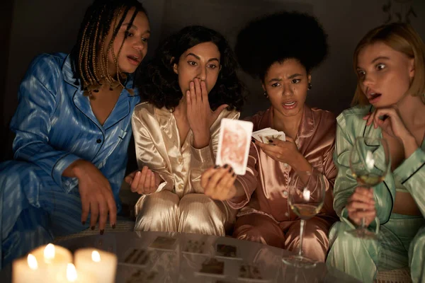 Weissagung, afrikanisch-amerikanische Frau im Pyjama hält verschwommene Tarotkarte in der Nähe schockierter multiethnischer Freundinnen, Weingläser und Kerzen während der Nacht der Mädchen zu Hause, Bindungszeit in Nachtwäsche — Stockfoto