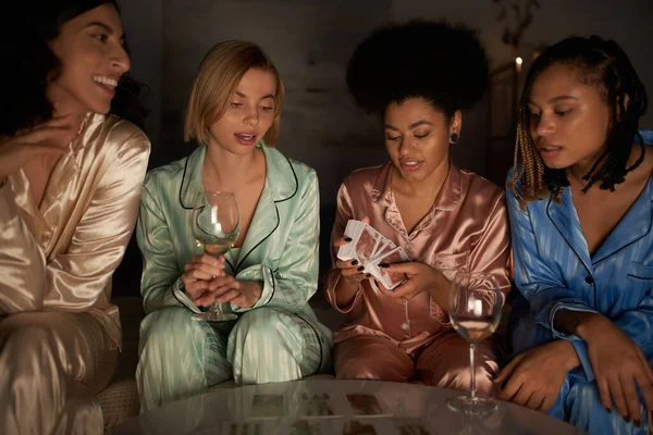Novias multiétnicas positivas en pijama sentadas cerca de un amigo afroamericano con cartas del tarot y copas de vino durante la noche de las niñas en casa, tiempo de vinculación en ropa de dormir cómoda, adivinación - foto de stock