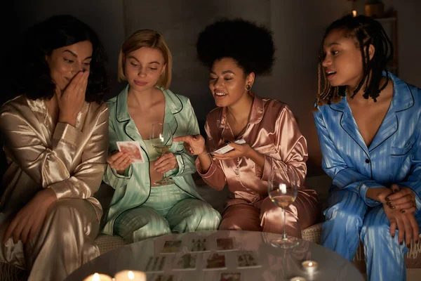 Positive multiethnische Freundinnen im farbenfrohen Pyjama, die Tarot-Karten in der Hand halten und sich bei Weingläsern und Kerzen während der Pyjama-Party zu Hause unterhalten, Bindungszeit in bequemer Nachtwäsche — Stockfoto