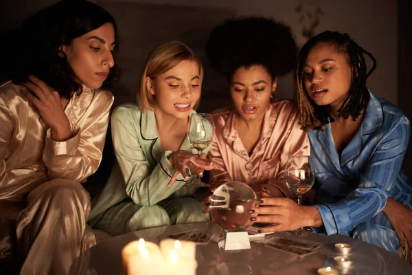 Petites amies multiethniques en pyjama coloré regardant boule de cristal près des verres de vin et des bougies sur la table pendant la nuit des filles à la maison, temps de liaison dans des vêtements de nuit confortables, divination — Photo de stock