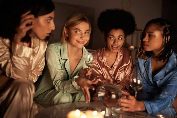 Femmes souriantes multiethniques en pyjama coloré regardant ami multiracial près de boule de cristal, verres à vin et bougies pendant la nuit des filles à la maison, temps de liaison dans des vêtements de nuit confortables, divination — Photo de stock