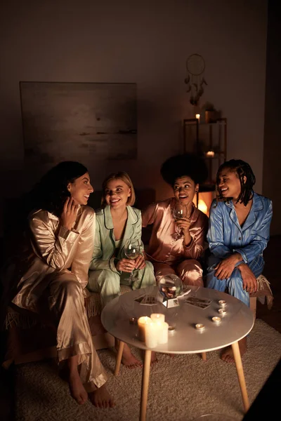 Mujeres alegres multiétnicas en pijama colorido hablando y sosteniendo copas de vino cerca de la bola de cristal y cartas del tarot durante la noche de las niñas en casa, tiempo de unión en ropa de dormir cómoda, adivinación - foto de stock