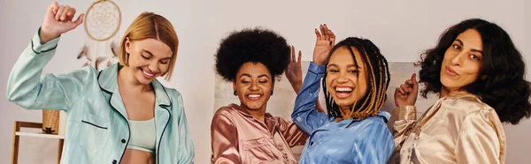 Positive multiethnische Freundinnen im farbenfrohen Pyjama tanzen und Spaß haben, während sie Zeit miteinander verbringen während Pyjama-Party zu Hause, Bindungszeit in bequemer Nachtwäsche, Schlummerparty, Banner — Stockfoto