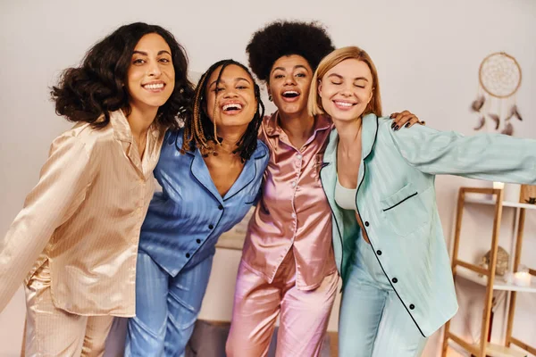 Lächelnde multiethnische Freundinnen im farbenfrohen Pyjama, die in die Kamera schauen, einander umarmen und bei der Schlummerparty zu Hause Spaß haben, Zeit in bequemer Nachtwäsche verbinden — Stockfoto
