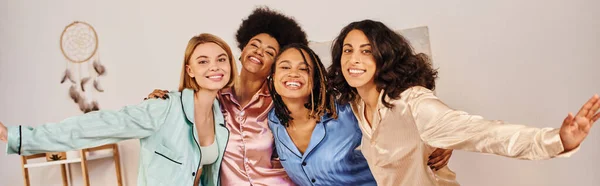 Positive multiethnische Freunde im bunten Pyjama, die sich umarmen und in die Kamera schauen, während sie zu Hause Spaß haben, Bindungszeit in bequemer Nachtwäsche, Schlummerparty, Banner — Stockfoto