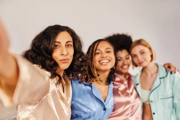 Multiracial woman in pajama looking at camera while standing near cheerful and blurred multiethnic girlfriends at home during slumber party, bonding time in comfortable sleepwear — Stock Photo