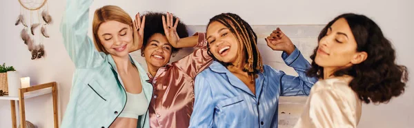 Petites amies multiethniques excitées et joyeuses en pyjama coloré dansant et s'amusant ensemble pendant la soirée pyjama à la maison, temps de liaison dans des vêtements de nuit confortables, bannière — Photo de stock