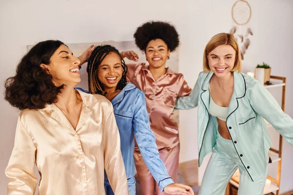 Mujer rubia positiva bailando cerca de novias multiétnicas en pijama colorido y mirando a la cámara juntos en casa durante la fiesta de pijamas, tiempo de unión en ropa de dormir cómoda - foto de stock