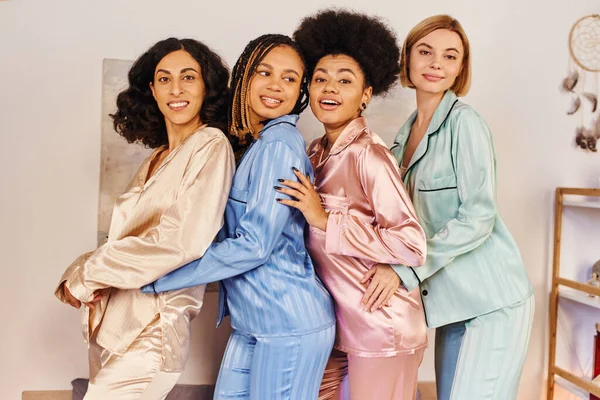 Fröhliche multikulturelle Freundinnen im bunten Pyjama, die in die Kamera schauen und sich beim Schlummerfest zu Hause umarmen, kulturelle Vielfalt, Bindungszeit in bequemer Nachtwäsche — Stockfoto