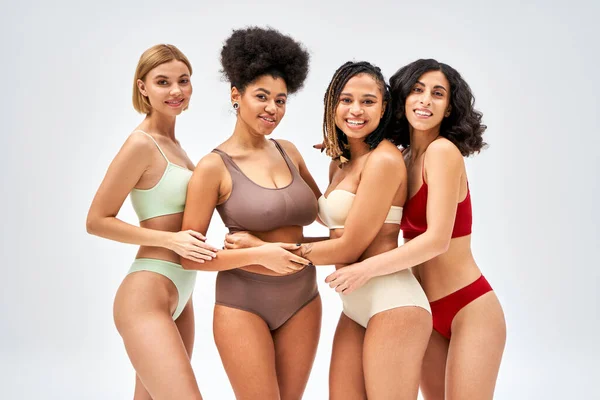 Cheerful multicultural girlfriends in colorful and modern lingerie hugging each other and standing isolated on grey, different body types and self-acceptance, multicultural representation — Stock Photo
