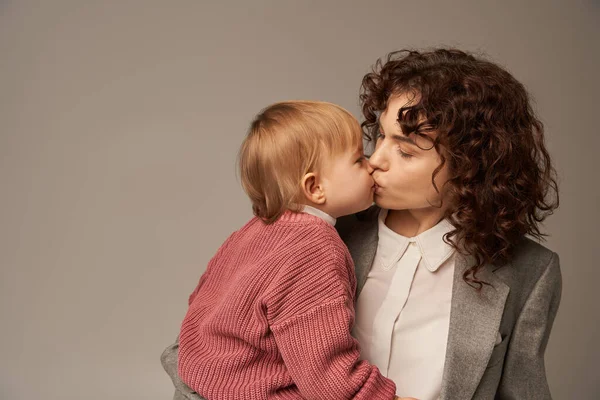 Work life balance concept, curly woman in suit kissing her daughter on grey background, loving mother, balanced lifestyle, modern working woman. modern parenting, career and family — Stock Photo