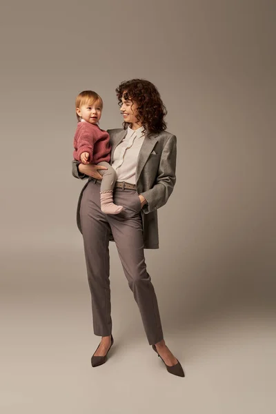 Quality time, work life balance concept, smiling woman in suit holding daughter and standing with hand in pocket on grey background, career and family, loving motherhood, full length — Stock Photo