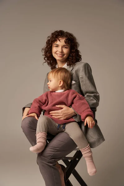 Balanced lifestyle, career and family, curly woman in suit sitting on chair with child on grey background, professional success, motherhood, multitasking, quality time, work life balance concept — Stock Photo
