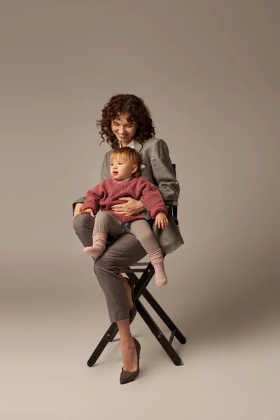 Збалансований спосіб життя, кар'єра та сім'я, кучерява жінка в костюмі сидить на стільці з дочкою на сірому фоні, професійний успіх, материнство, багатозадачність, якісний час, концепція балансу життя — стокове фото