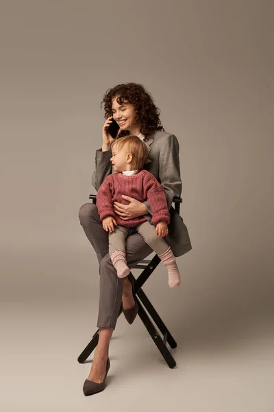Estilo de vida equilibrado, telefonema, mulher de negócios feliz falando no smartphone e sentado na cadeira com criança em fundo cinza, maternidade, multitarefa, tempo de qualidade, conceito de equilíbrio de vida no trabalho — Fotografia de Stock